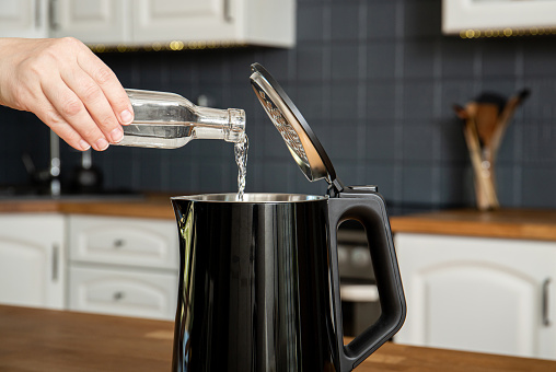 Woman pouring natural destilled acid white vinegar in electric kettle to remove boil away the limescale. Descaling a kettle, remove scale concept. Home kitchen background.