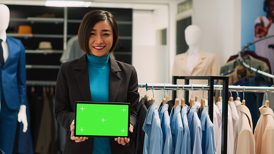 Female asian woman using green screen on digital tablet, holding blank chroma key display in department store. Young shop assistant having isolated mockup and copyspace template.