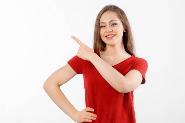 istock Smiling lovely girl points left at promo banner, shows advertisement on blank white space. Young brunette female looking happy by promotion offer, stands over white background 1475239095