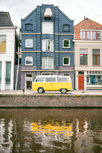 Classic Volkswagen Transporter T2 campervan, parked on a residential street next to a canal in Groningen, Netherlands. The bright yellow van stands out from the dark house in the background.