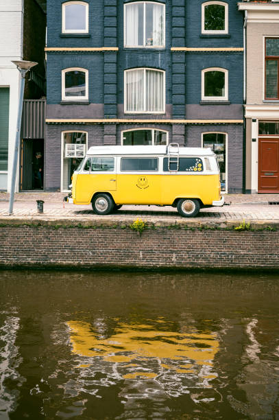 Classic Volkswagen Transporter T2 campervan, parked on a residential street next to a canal in Groningen, Netherlands. The bright yellow van stands out from the dark house in the background.