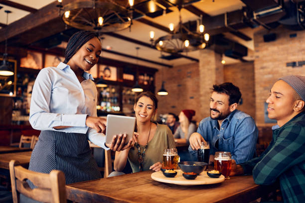 istock Happy black waitress using touchpad while suggesting order from menu to her customers in a bar. 1475239941