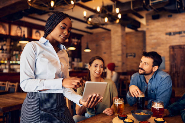 istock Young black waitress recommending order on touchpad while serving guests in a pub. 1475239956
