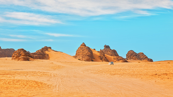 Typical desert landscape in Alula, Saudi Arabia, sand with some mountains, small offroad vehicle, local man and camels at distance.
