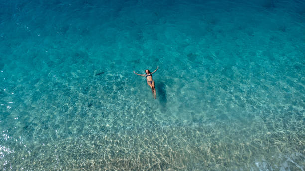 Aerial view of beautiful happy woman in swimsuit laying in the shallow sea water enjoying beach and soft turquoise ocean wave. Tropical sea in summer season on Egremni beach on Lefkada island. Aerial view of beautiful happy woman in swimsuit laying in the shallow sea water enjoying beach and soft turquoise ocean wave. Tropical sea in summer season on Egremni beach on Lefkada island. egremni beach lefkada island greece stock pictures, royalty-free photos & images