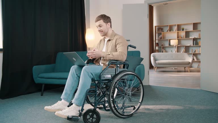 Cheerful disabled caucasian man on wheelchair using laptop in living room at home