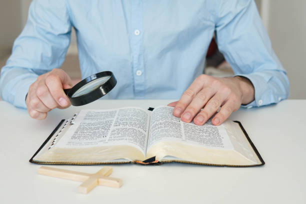 istock Christian catholic man with magnifying glass searching for answers in old Holy Bible. Loupe on a Bible. 1475253473
