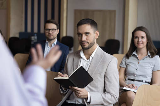 Unrecognizable presenter make speech standing in front of participants noting received information looking interested, motivated to develop their skills during educational business seminar in office