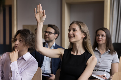 Smiling woman employee, educational seminar participant raise arm to ask question engaged in teambuilding activity during meeting or briefing in conference hall. Interview or workshop, opinion, vote