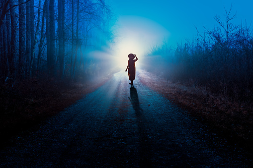A silhouette of a woman in a dress and hat on a foggy night forest country road walking into a bright car head lights.