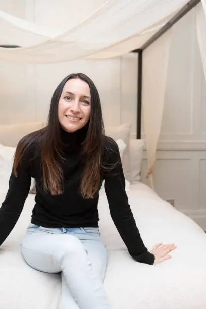 Relaxed and calm portrait of a woman in a bedroom. See is wearing a black polo neck jumper and jeans.