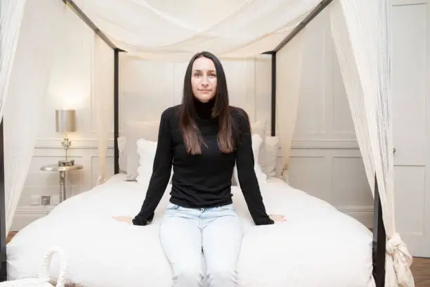 Relaxed and calm portrait of a woman in a bedroom. See is wearing a black polo neck jumper and jeans.