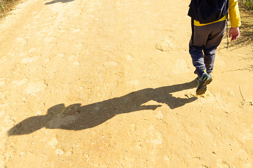 Boy on his back walking with backpack practicing hiking. reflection of his shadow on the dirt road. Copy space
