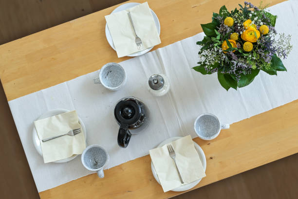 Wooden table with three plates, cups, napkins, coffee pot and a flower bouquet on a white table runner, high angle view from above, selected focus stock photo