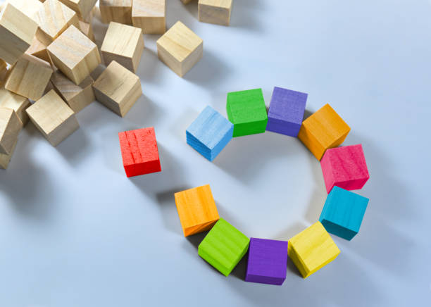Cubes in different colors together form a circle with a gap, one is red and stands separate, heap of natural wooden cubes in the corner, light blue background, copy space