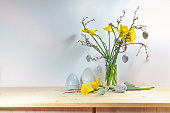 Spring decoration with a bouquet of daffodils and branches in a glass vase and artificial blue gray Easter eggs on a wooden sideboard against a light wall, copy space