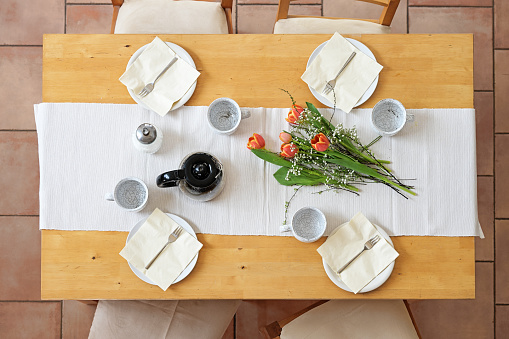 Setting for an afternoon coffee break with four plates, cups, napkins, coffee pot and tulip flowers on a white table runner on a wooden table, top view from above, selected focus