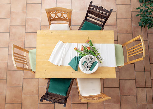 Preparation for dinner with table runner, white plates, cutlery, green napkins and some tulip flowers, six different wooden chairs, tiled terracotta floor, top view from above, selected focus