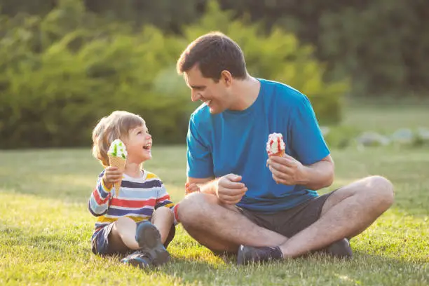 Father and son eating ice cream in waffel cone and fave fun. Laughing family sitting on grass in city park. Happy fathers day.