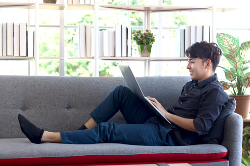 Young handsome Asian man in black shirt typing on keyboard laptop computer while lying down comfortably on sofa couch in his apartment, happy relaxing freelancer working online in living room.