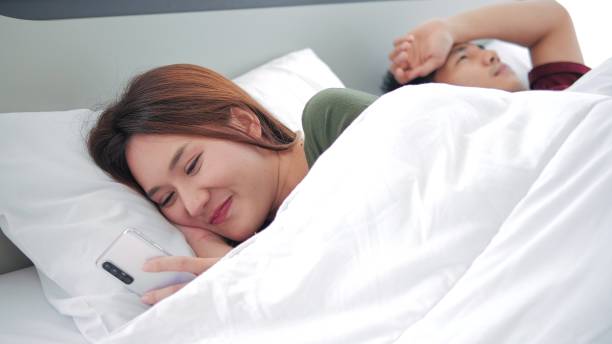 幸せな笑顔のアジア人女性は、恋人からの電話でメッセージを読んでいる男性に背を向け、彼女の隣に横たわる心配なボーイフレンド、浮気と不貞のコンセプト - sleeping lying on back couple bed ストックフォトと画像
