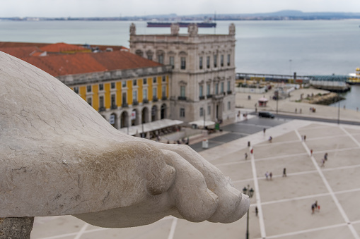 Lisbon, Portugal panoramic landscape defocused view of Commerce square, Praca do Comercio, with front ground focus on art detail marble leg on top of 1875 triumphal arch Arco da Rua Augusta.