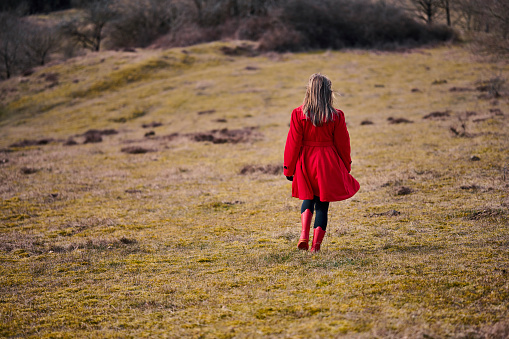 Outdoors portrait of woman in red. Rear view of walking white woman with long hair wearing warm clothing. Walking in meadow with red rubber boots