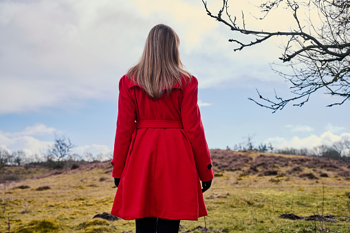 Outdoors portrait of woman in red. Rear view of walking white woman with long hair wearing warm clothing. Walking in meadow with red rubber boots