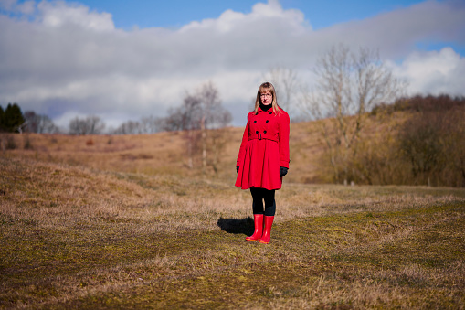 Outdoors portrait of woman in red. Rear view of walking white woman with long hair wearing warm clothing. Walking in meadow with red rubber boots