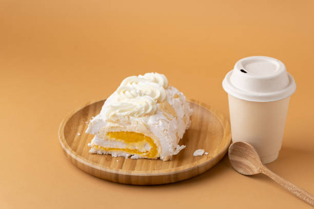 istock Meringue roll filled with mango puree on the brown background. Meringue roulade with yellow fruit filling in the bamboo wooden plate next to beige paper cup of coffee or tea. Sweet pastry concept 1475276443