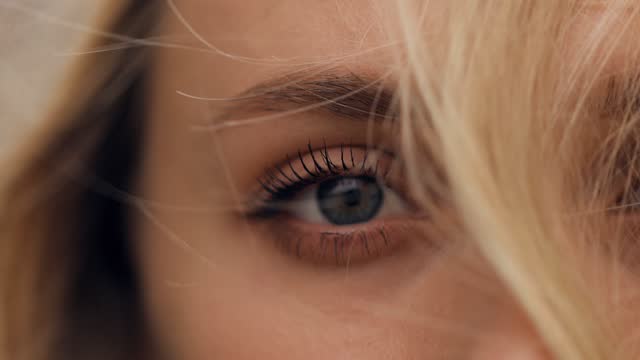 close-up macro of the blue eye of a blond-haired woman, her hair blowing in the wind