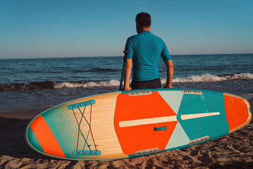 Rear view of a man, paddleboard surfer, enjoying the sea view and relaxing on the beach.