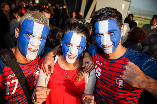 Two men and a woman in a crowd of spectators going on a sports match. Soccer, american football, spectators excited outside the stadium. Face painting in club colors. Excitment on their faces.