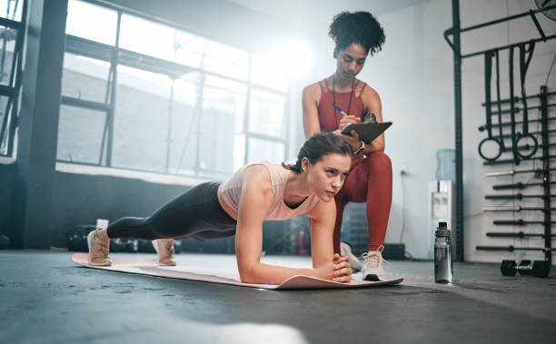 personal trainer, fitness e appunti con una donna di colore che allena un cliente in una palestra durante il suo allenamento. salute, esercizio fisico o allenamento e un'atleta che fa un plank con il suo allenatore che scrive note - endurance sport foto e immagini stock