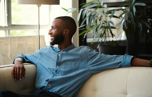 Portrait, relax and smile of black man on sofa in home in comfortable living room. Happy young male from South Africa relaxing and sitting on couch in lounge of he's  house.