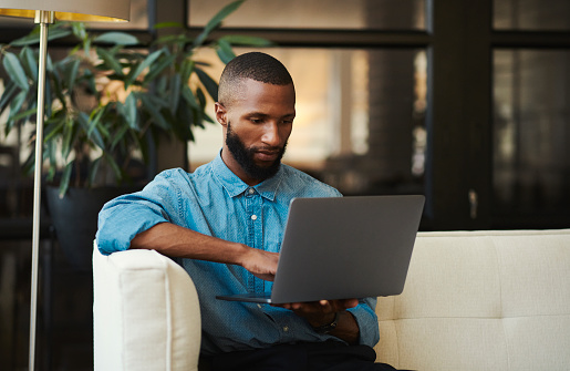 Freelancer with a laptop in the living room with a black man doing remote work on he's home sofa. Computer, small business and young male freelancer or entrepreneur working at home