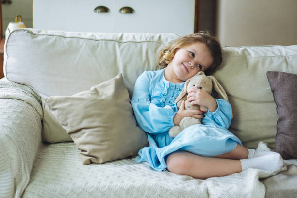bambina carina in vestito blu sul divano di casa con il suo giocattolo preferito, il coniglietto di peluche. concetto di bambini che giocano - 4 of a kind foto e immagini stock