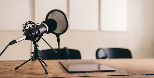 Still life shot of a microphone with a pop shield placed on a table alongside a digital tablet. Podcast recording equipment set in a home studio.