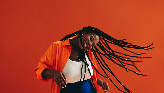 Vibrant young woman flipping her hair playfully while standing in a studio. Excited woman dancing and celebrating against an orange background. Happy young woman having fun with her dreadlocks.