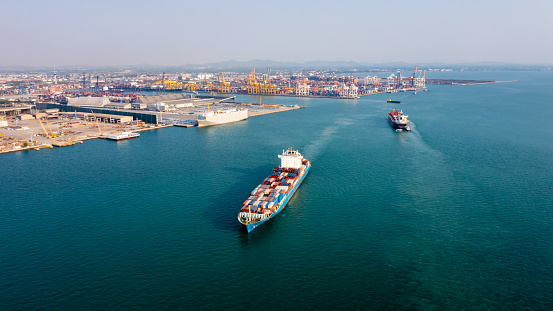 front view on a tugboat towing a large cargo container ship with piled up cargo containers on deck into harbor.