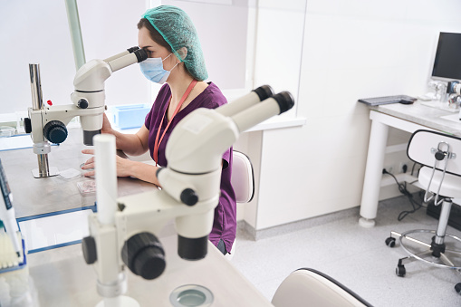Woman laboratory assistant looks into the eyepiece of powerful microscope, at her workplace there is modern equipment for vitrification procedure