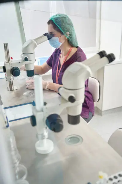 Photo of Woman in overalls looks into eyepiece of a powerful microscope