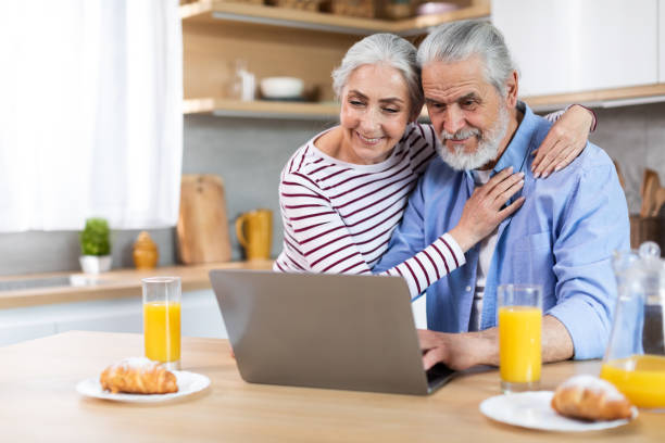 smiling senior couple using laptop in kitchen while having breakfast together - two parent family indoors home interior domestic kitchen imagens e fotografias de stock