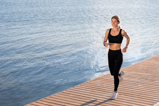 Morning Jogging. Beautiful sporty middle aged woman running outdoors on wooden pier along river, smiling fit female runner in sportswear training outside, enjoying healthy lifestyle, copy space