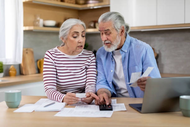 senior couple sitting at table with papers and bills and using calculator - two parent family indoors home interior domestic kitchen imagens e fotografias de stock