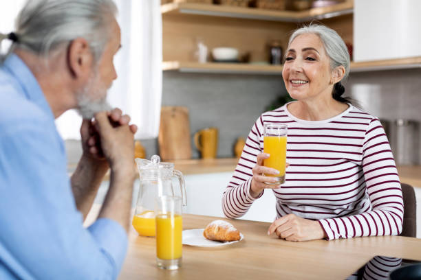 happy senior woman drinking juice while having breakfast with husband in kitchen - two parent family indoors home interior domestic kitchen imagens e fotografias de stock