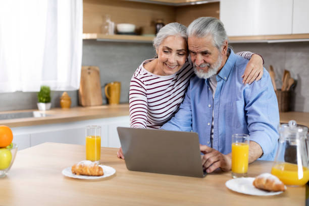 happy elderly spouses spending time with laptop in kitchen - two parent family indoors home interior domestic kitchen imagens e fotografias de stock