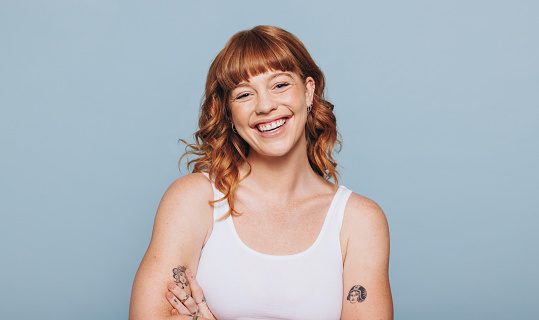 Happy red haired woman smiling at the camera in a studio. Portrait of a young caucasian woman standing in a white tank top.