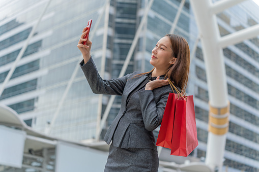 Photo of a young asian businesswoman in grey office suit using her mobile phone walking happily across a central business district with red shopping paper bag in hand