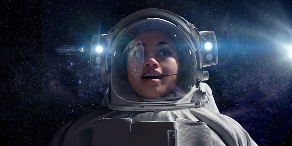 A close up head and shoulders image of a young mixed race female astronaut on a spacewalk, looking with wide eyes and open mouth in awe at earth which is reflected in her helmet's visor. She is wearing a plain white space suit and her helmet has lights which are on.